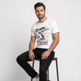 Man wearing a white t-shirt with a graphic design sitting on a stool against a plain background