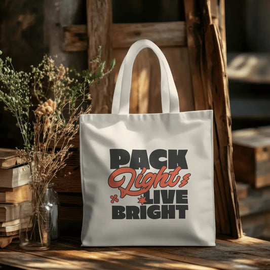 Tote bag with 'Pack Light Live Bright' text on a wooden surface with books and plants.