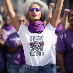 Woman with raised fists wearing a white t-shirt with a black and red design and purple sunglasses, surrounded by people in a protest or rally setting.