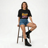 Woman sitting on a stool wearing a black t-shirt with colorful text and denim shorts.