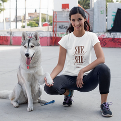 Woman squatting with a dog on a concrete surface, wearing a 'SHE' t-shirt.