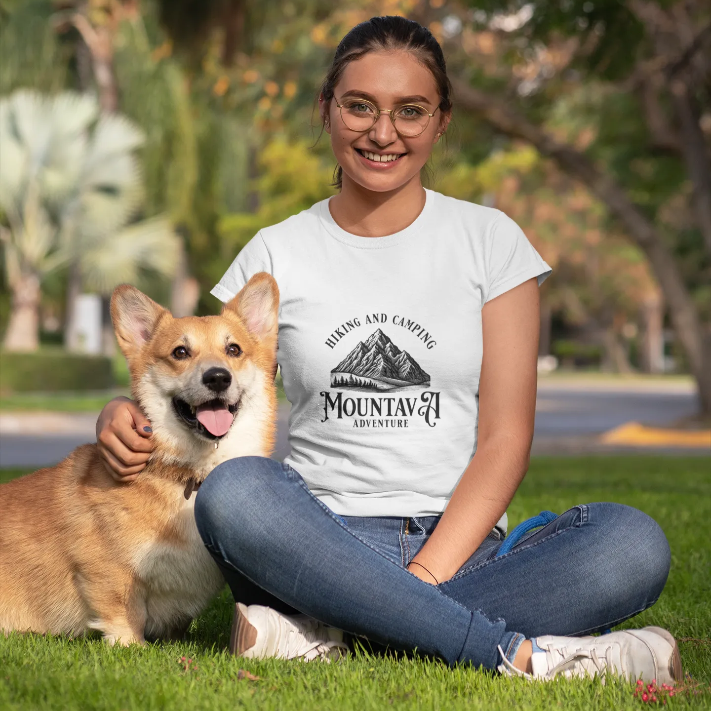 Woman sitting on grass with a dog, wearing a white t-shirt with a mountain design.