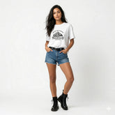 Woman wearing a white t-shirt with a mountain design and text, paired with denim shorts and black boots on a white background.