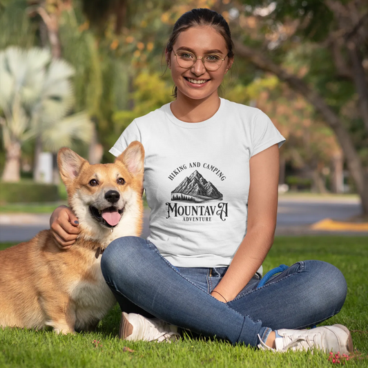 Woman sitting on grass with a dog, wearing a white t-shirt with a mountain design.