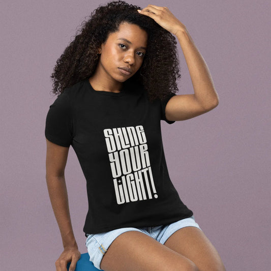 Woman wearing a black t-shirt with 'Shine Your Light!' text, sitting on a blue box against a purple background