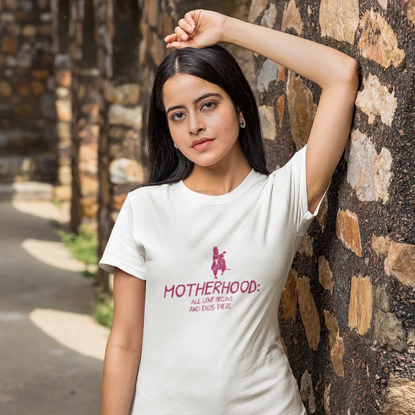 Woman wearing a white t-shirt with a graphic and text, standing against a stone wall.