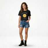 Woman wearing a black t-shirt with a sun graphic and text, standing against a light-colored wall.