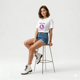 Woman sitting on a stool wearing a white t-shirt with a graphic design, denim shorts, and white boots on a light gray background.