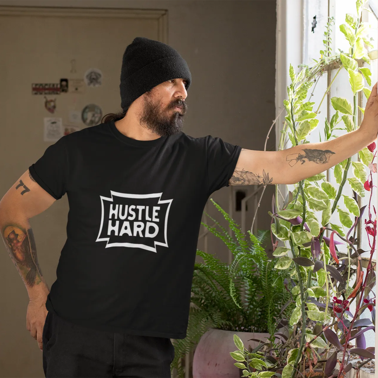 Man wearing a black t-shirt with 'Hustle Hard' text, standing indoors with plants.
