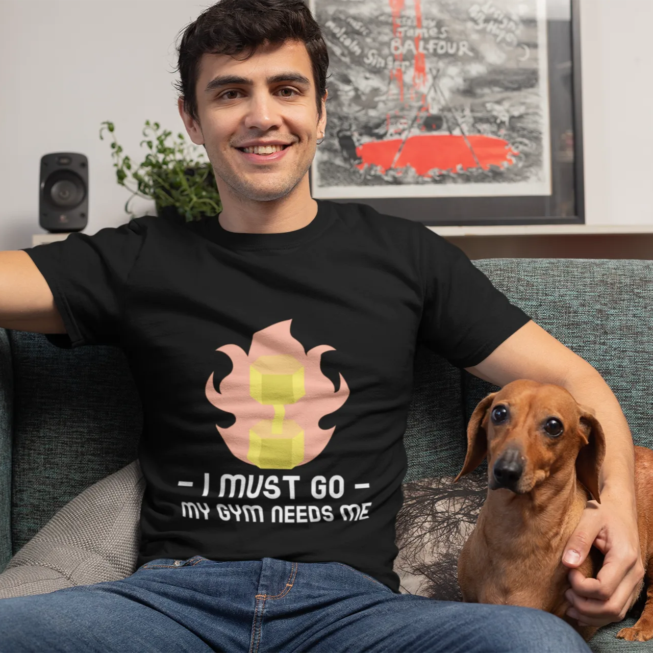 Man sitting on a couch with a dog, wearing a black t-shirt with a graphic design and text.
