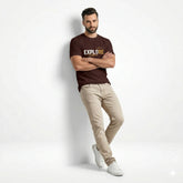 Man wearing a brown t-shirt with 'EXPLORE' text, beige pants, and white shoes on a light gray background
