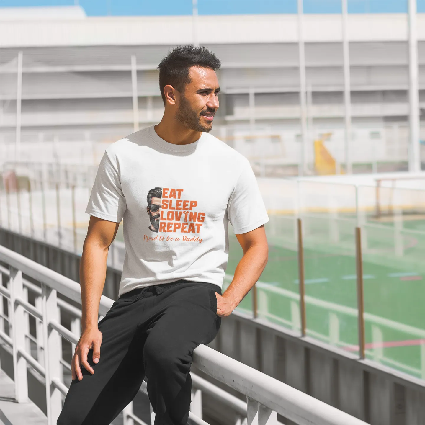 Man wearing a white t-shirt with text and graphics, standing outdoors near a sports field.