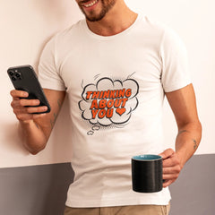 Man wearing a white t-shirt with 'Thinking About You' text, holding a phone and a mug.
