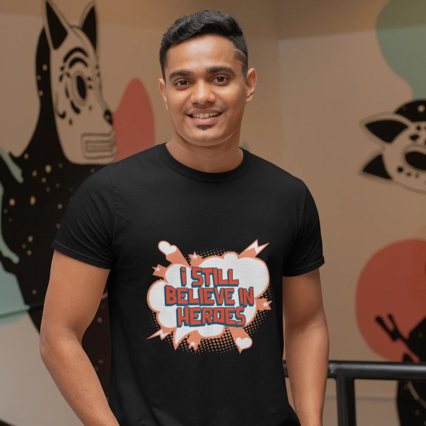 Man wearing a black t-shirt with 'I Still Believe in Heroes' text, standing in front of colorful wall art.