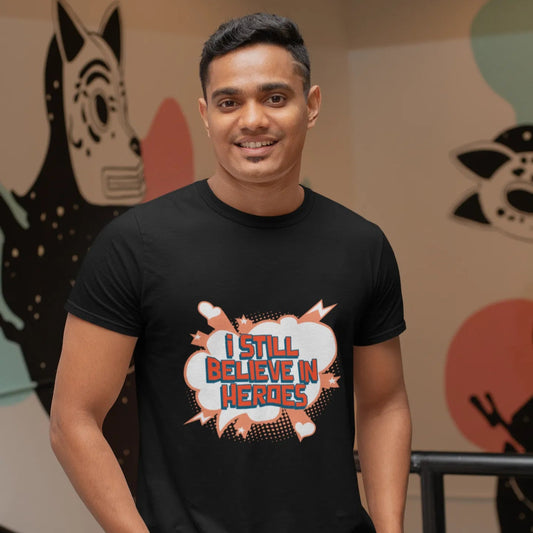 Man wearing a black t-shirt with 'I Still Believe in Heroes' text, standing in front of colorful wall art.