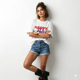 Woman wearing a white t-shirt with 'Happy Holi' text, denim shorts, and black boots on a light background