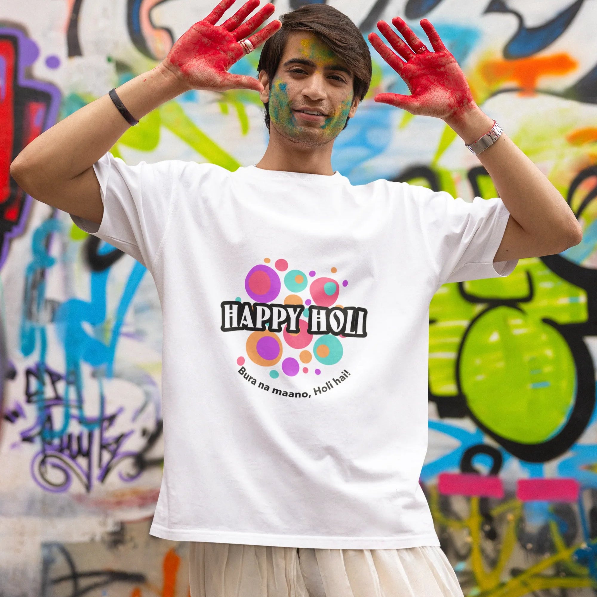 Person wearing a 'Happy Holi' t-shirt with colorful paint on hands against a graffiti-covered wall.