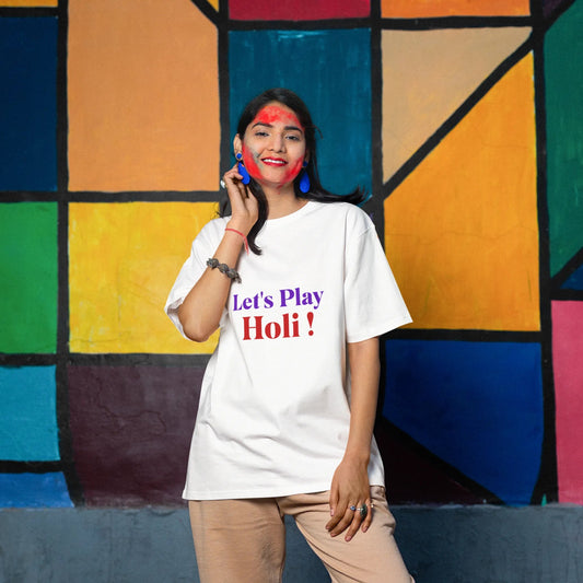 Person wearing a white t-shirt with 'Let's Play Holi!' text in front of a colorful geometric-patterned wall.