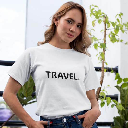 Woman wearing a white t-shirt with 'TRAVEL.' text, standing outdoors with plants in the background.