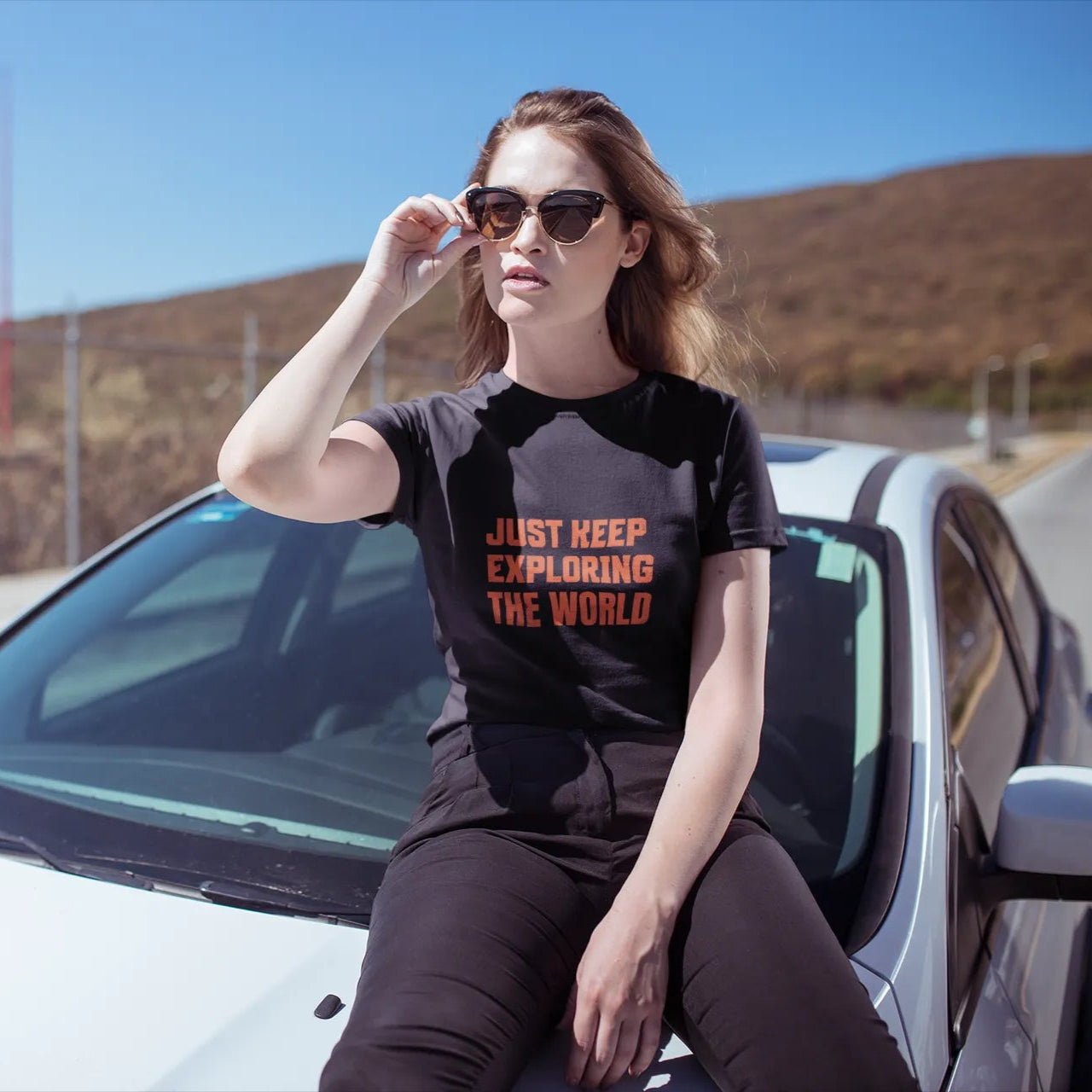 Woman sitting on a car wearing a t-shirt with text, with a scenic background