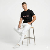 Man sitting on a stool wearing a black t-shirt with 'MUSAFIR!!' text and white pants.
