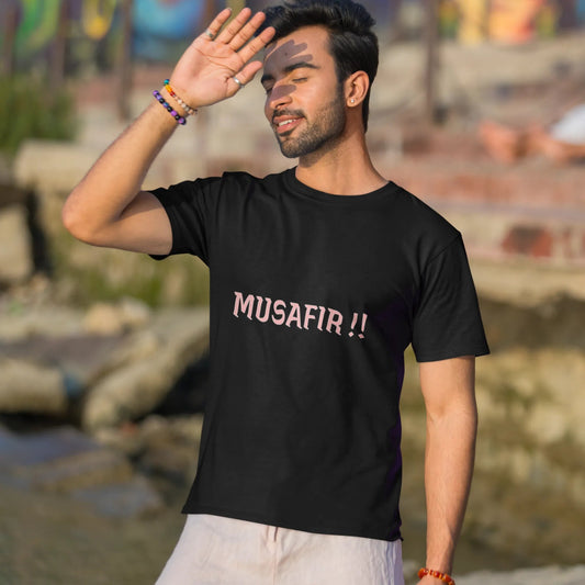 Man wearing a black t-shirt with 'MUSAFIR!!' text, standing outdoors with a colorful mural in the background.