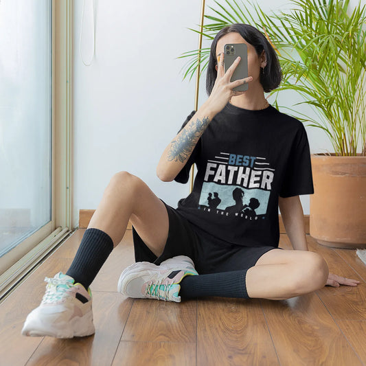 Person sitting on the floor wearing a black t-shirt with 'Best Father' graphic, taking a mirror selfie.