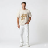 Man wearing a beige t-shirt with text and white pants on a light gray background
