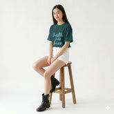 Person wearing a teal t-shirt with 'Faith Over Fear' text, sitting on a wooden stool against a white background.