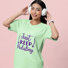 Woman wearing a light green t-shirt with 'Just Keep Pedaling' text, white headphones, and blue jeans on a pink background.