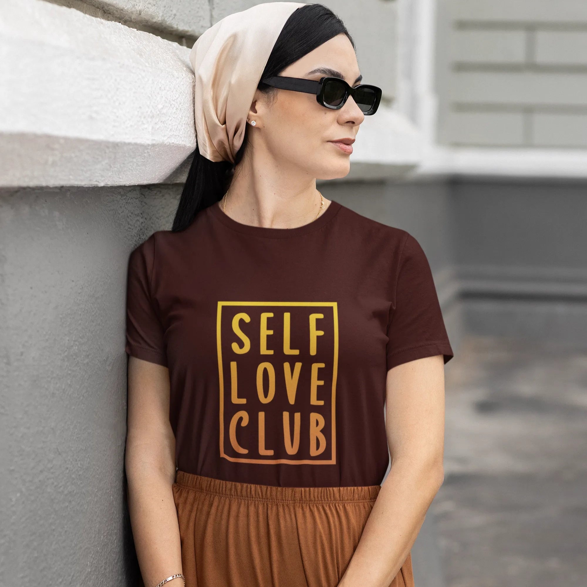 Woman wearing a brown t-shirt with 'SELF LOVE CLUB' text, standing against a gray wall.