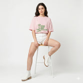 Woman sitting on a stool wearing a pink t-shirt with text and white shorts on a light gray background