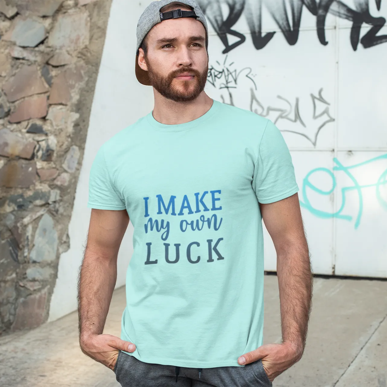 Man wearing a light blue t-shirt with 'I Make My Own Luck' text, standing against a graffiti-covered wall.