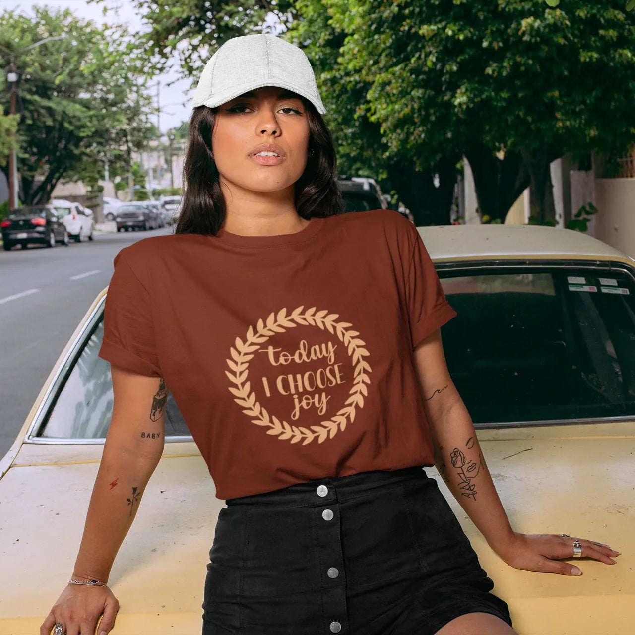 Woman wearing a brown t-shirt with text standing next to a yellow car on a street.