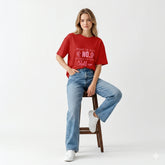 Woman wearing a red t-shirt and blue jeans sitting on a stool against a white background