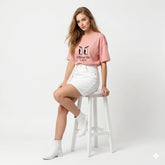 Woman wearing a pink graphic t-shirt and white shorts sitting on a stool against a white background