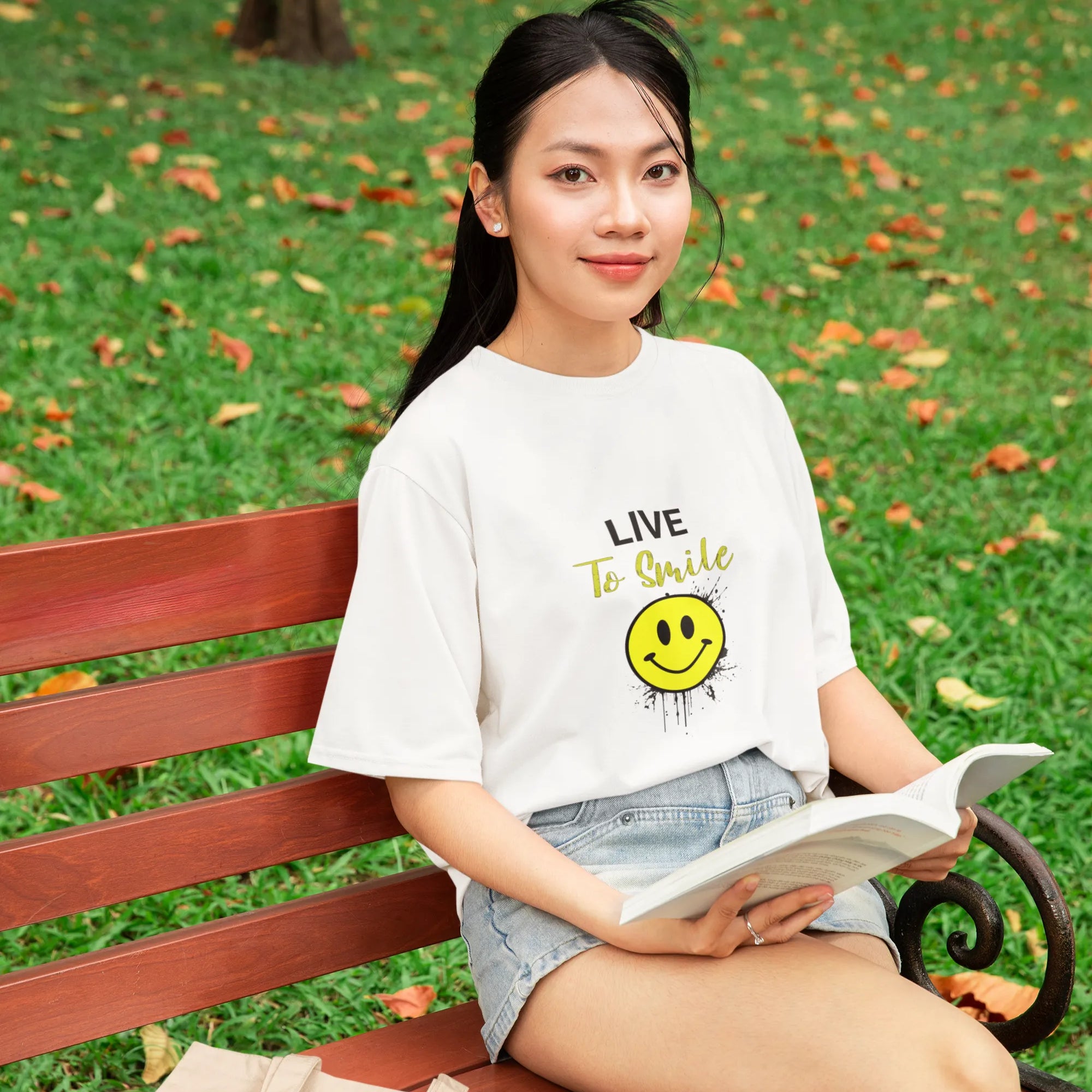 Woman sitting on a park bench reading a book, wearing a white t-shirt with a smiley face design.