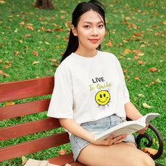 Woman sitting on a park bench reading a book, wearing a white t-shirt with a smiley face design.