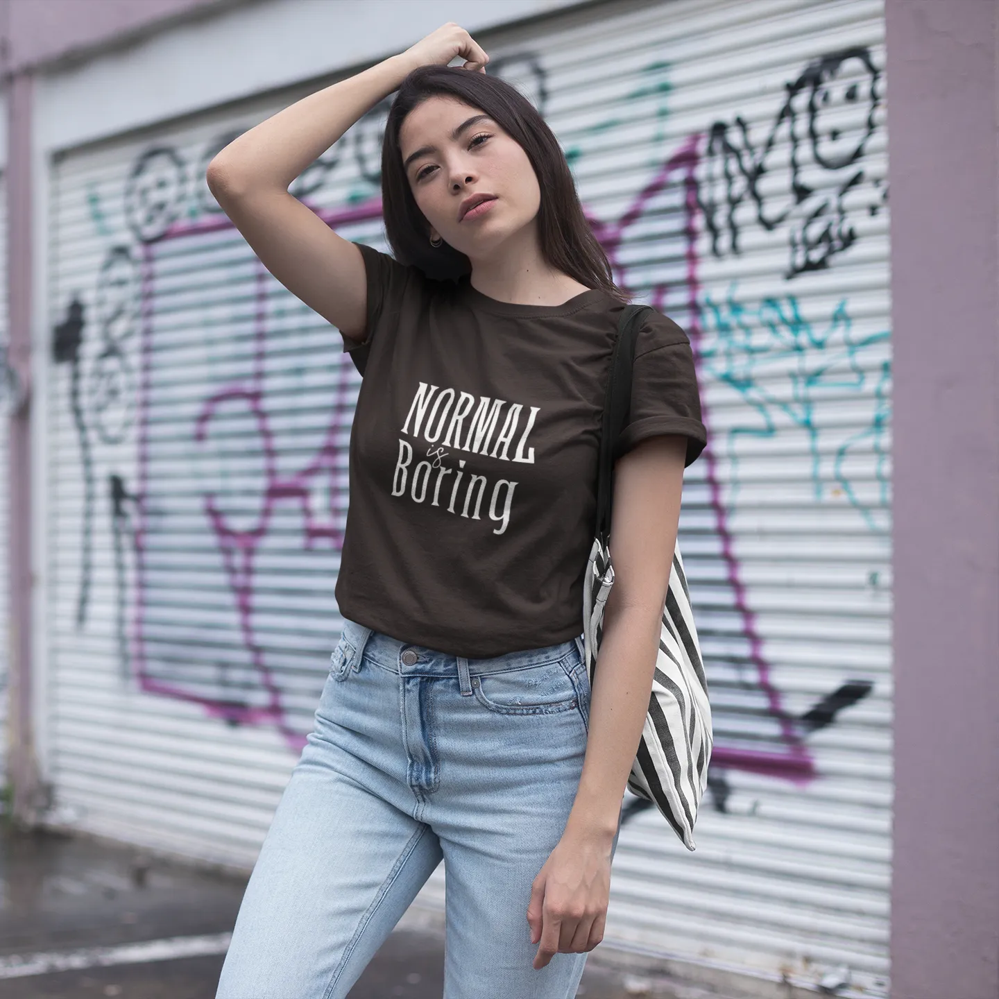 Woman wearing a brown t-shirt with 'Normal is Boring' text in front of a graffiti-covered wall.