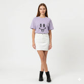 Woman wearing a light purple t-shirt with a smiley face design and a white skirt on a white background
