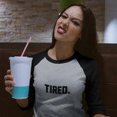 Woman wearing a 'TIRED.' shirt holding a drink in a casual setting