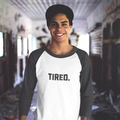 Person wearing a white and black baseball shirt with 'TIRED.' text in an indoor setting.