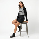 Woman sitting on a stool wearing a black and gray long-sleeve shirt with 'VIBE' text, black shorts, and black boots.