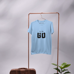 Light blue t-shirt with 'Let's Go' text on a hanger, next to a brown suitcase and potted plant.