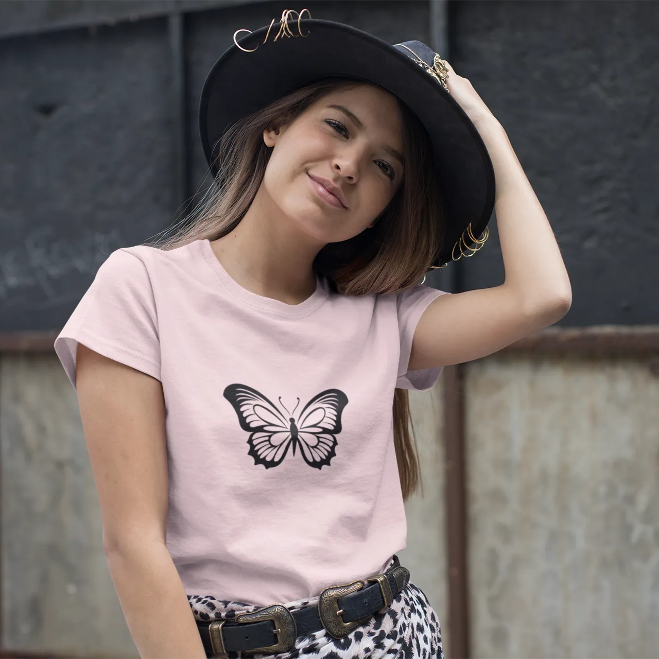 Woman wearing a pink t-shirt with a butterfly design and a black hat against a textured wall.
