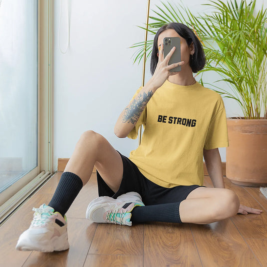 Person sitting on the floor wearing a yellow 'BE STRONG' t-shirt, taking a mirror selfie.