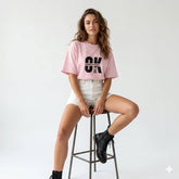 Woman wearing a pink t-shirt with text, sitting on a stool against a white background