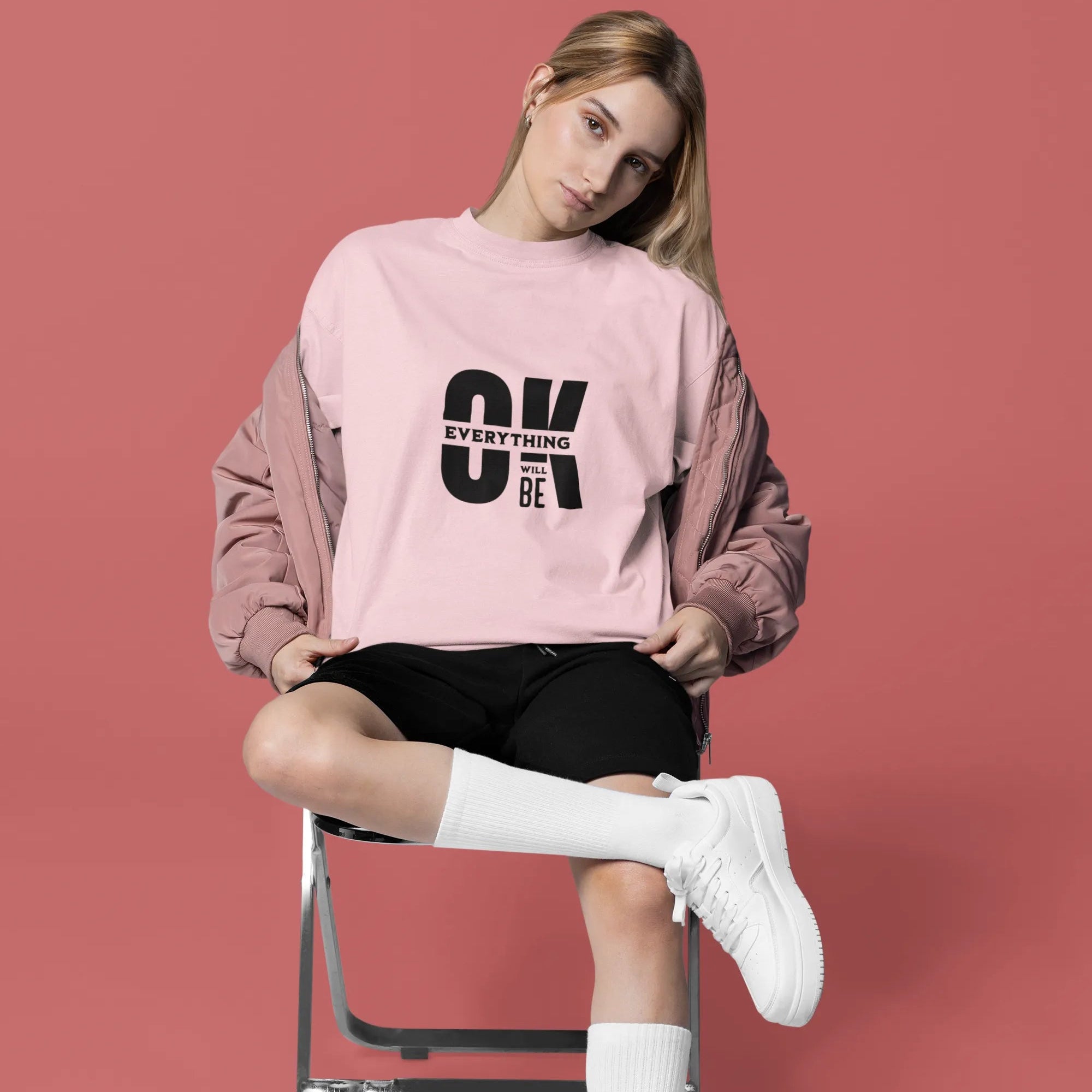 Woman sitting on a chair wearing a pink sweatshirt with text, black shorts, white socks, and white sneakers on a red background.