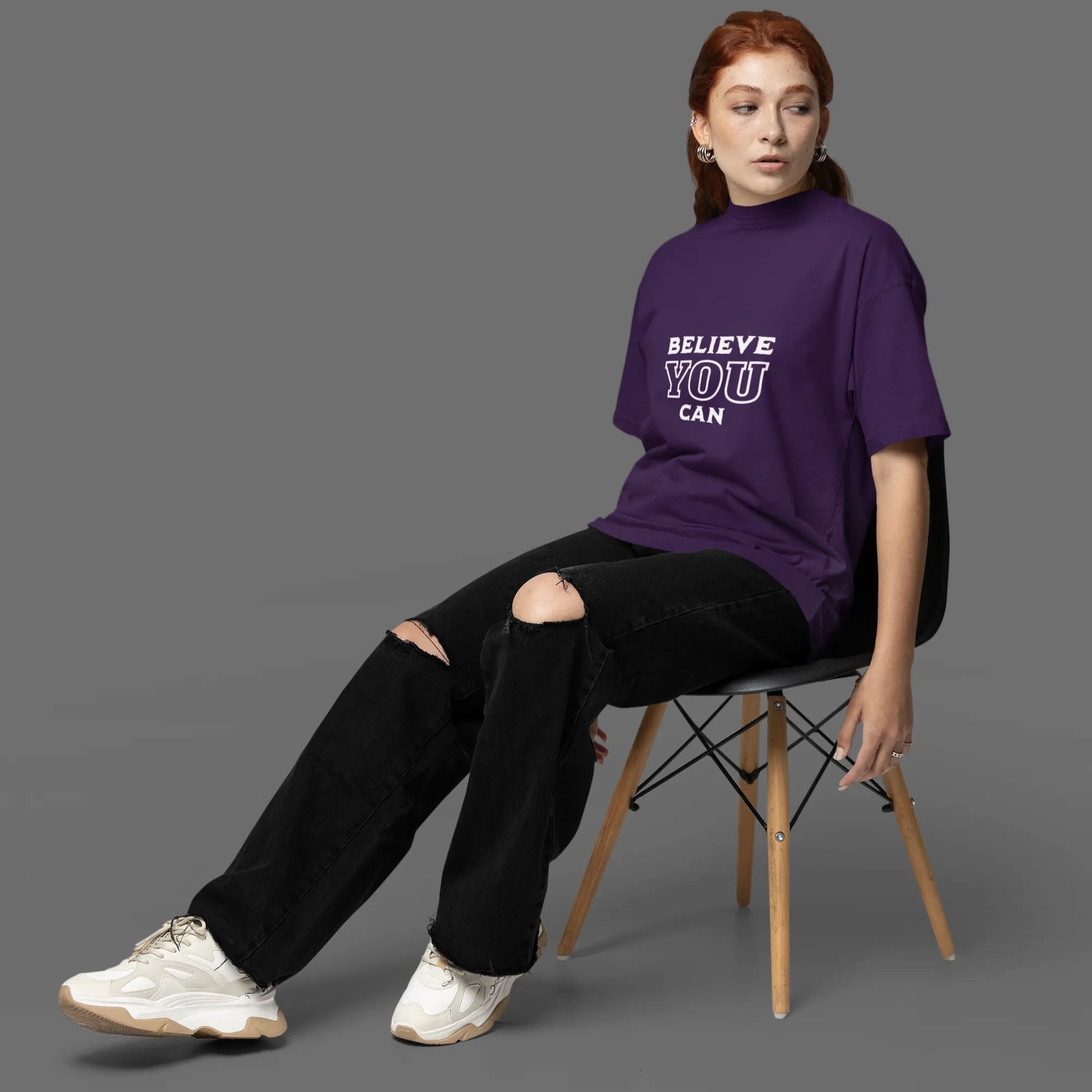 Person wearing a purple t-shirt with 'BELIEVE YOU CAN' text, sitting on a chair against a gray background