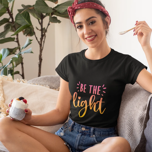 Woman wearing a black t-shirt with 'Be the light' text, sitting on a couch holding a mug.